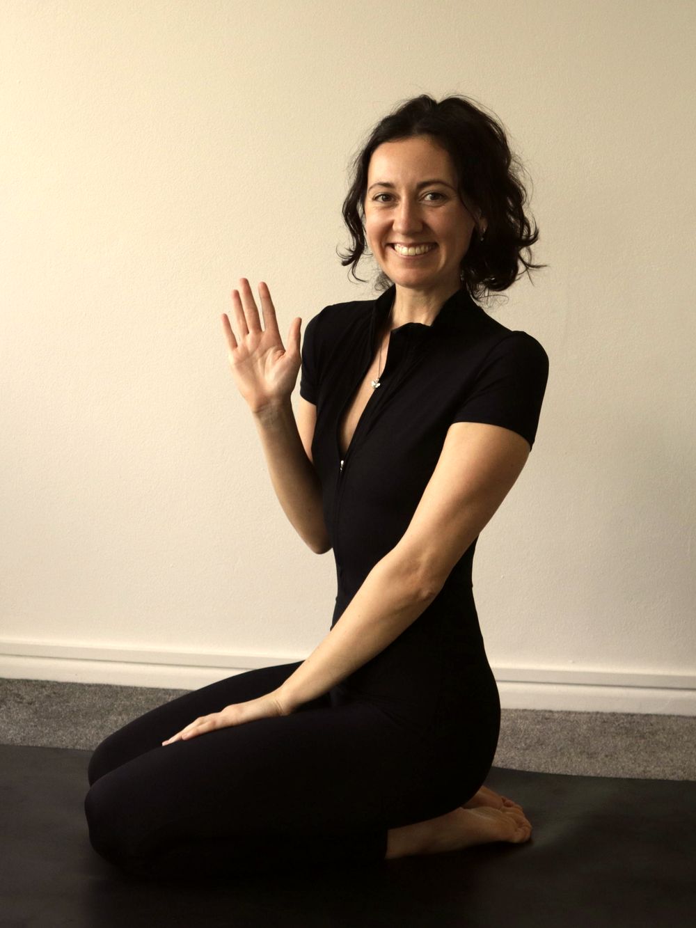 Issy Luck kneeling on a yoga mat and waving to camera at the start of a class